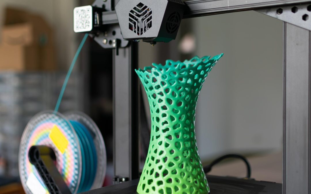 A green, lattice-patterned vase being 3D printed on a desktop 3D printer with filament spool in background.